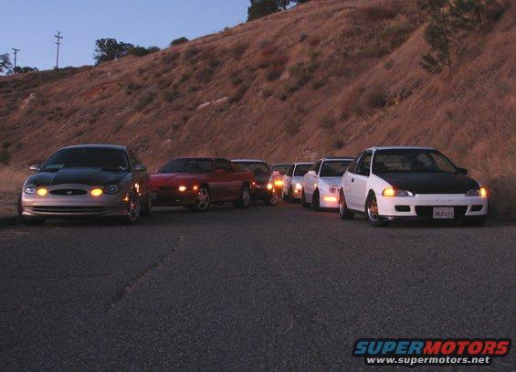 Several cars parked in a line on a paved road with a hillside in the background
