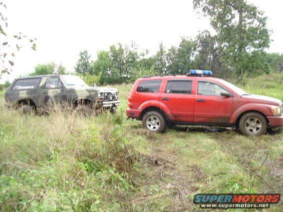 Camouflaged Ford Bronco stuck in mud, red SUV with light bar nearby