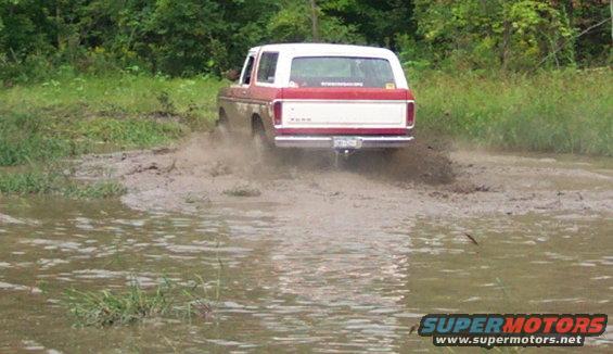 Red and white Ford Bronco driving through mud, rear view