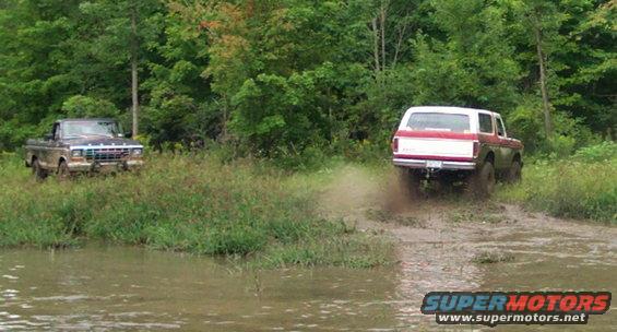 Red and white 1979 Ford Bronco driving through mud, with a pickup truck nearby