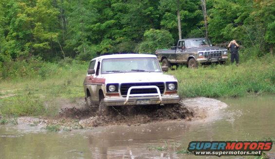 White 1978 Ford Bronco driving through mud, with a black Ford F-Series truck behind it