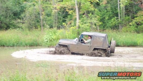 Mud-splattered Jeep CJ-8 Scrambler with a soft top driving through a muddy field