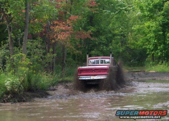 Red pickup truck with dual exhaust pipes driving through a muddy puddle, kicking up water and mud