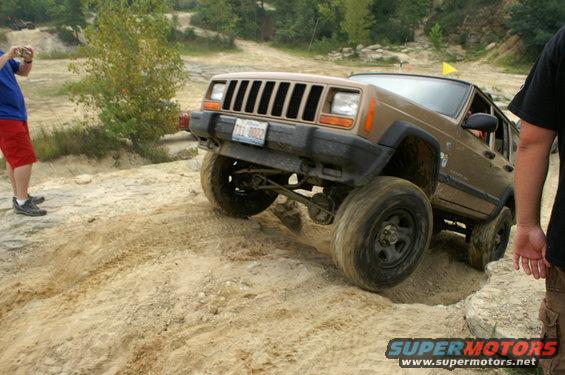 Brown Jeep Cherokee XJ climbing sandy hill, driver's side front wheel off ground. Person in blue shirt and red shorts taking photo