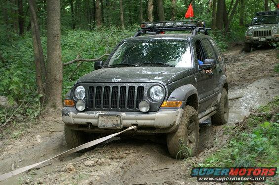 Dark gray Jeep Liberty on a muddy trail, being pulled by a strap. Another Jeep is visible behind it in the woods