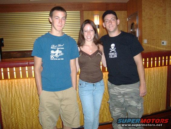 Three people stand in front of a textured wall. The person on the left wears a blue t-shirt and tan shorts. The person in the middle wears a brown tank top and jeans. The person on the right wears a black t-shirt and camouflage pants