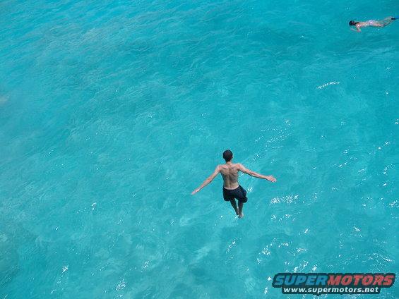 Man jumping into blue ocean water with another person swimming in distance. Supermotors.net logo