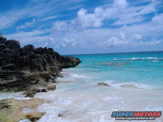 Beach with rocky outcroppings and turquoise water under a cloudy blue sky. "SUPERMOTORS" logo in lower right