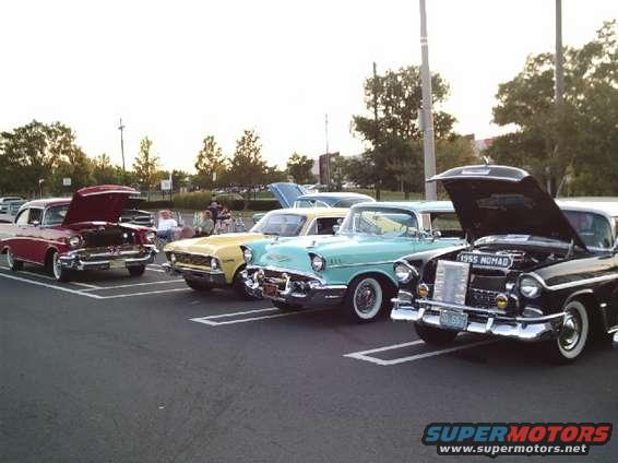 Several classic cars parked in a lot, hoods up on some. One has a '1955 NOMAD' badge