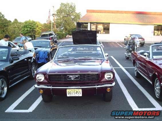 Maroon 1965 Ford Mustang convertible parked in a lot, with another Mustang and a motorcycle nearby