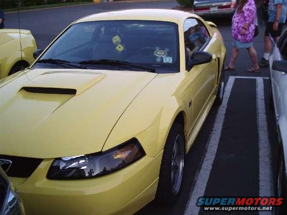 Yellow Ford Mustang parked in a lot, front quarter view. Dice hanging from rearview mirror