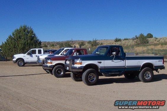 Three pickup trucks parked on a dirt lot under a clear blue sky. The closest truck is a two-tone green and white Ford F-150