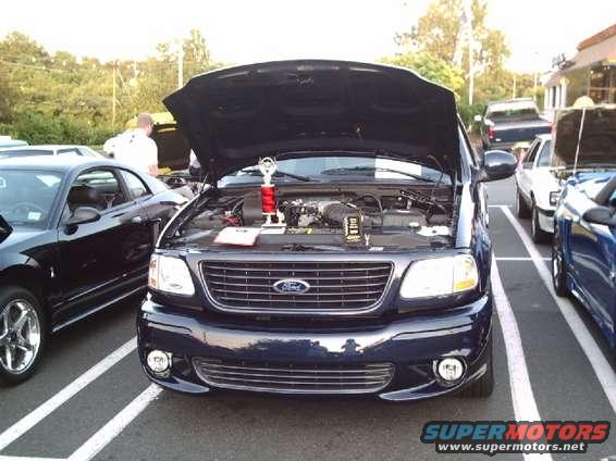 Dark blue Ford F-150 SVT Lightning with hood open, trophy on engine. Parked next to a black car