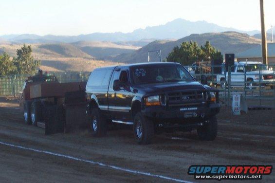 Dark blue Ford F-Series truck with a cap, pulling a sled. Another white truck is visible in the background