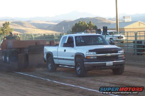 White Chevrolet Silverado pickup truck pulling a sled at a truck pull event. Another white truck is parked in the background