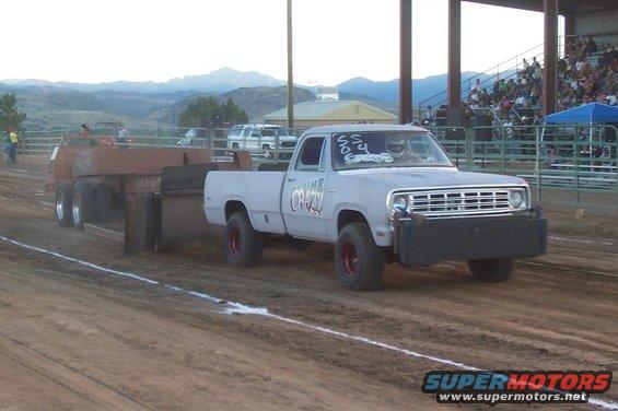 Light blue pickup truck with red wheels and a black bumper pulls a trailer in a dirt track event