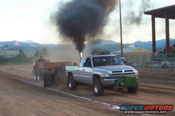Silver Dodge Ram pickup truck with large exhaust stack emitting black smoke, pulling a sled in a competition. Green front bumper/weight