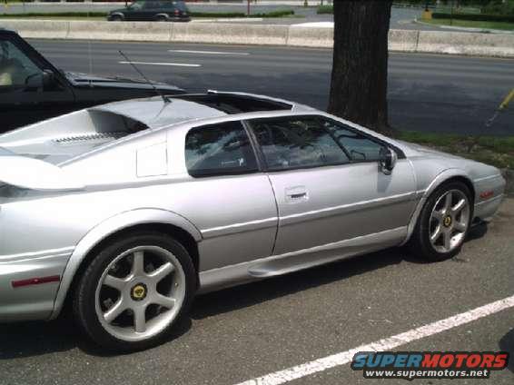 Silver sports car with a rear spoiler, parked on asphalt. A black SUV is partially visible on the left