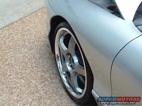 Silver car fender and five-spoke alloy wheel with a chrome lip, parked on a pebbled surface