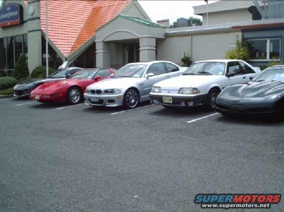 Several sports cars, including a white Ford Mustang, a red Chevrolet Corvette, and a silver BMW, are parked in a lot outside a building