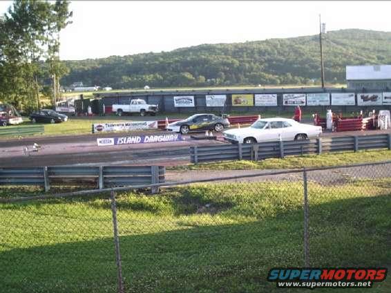White coupe drag racing on a track, side view. Appears to be from the 1970s