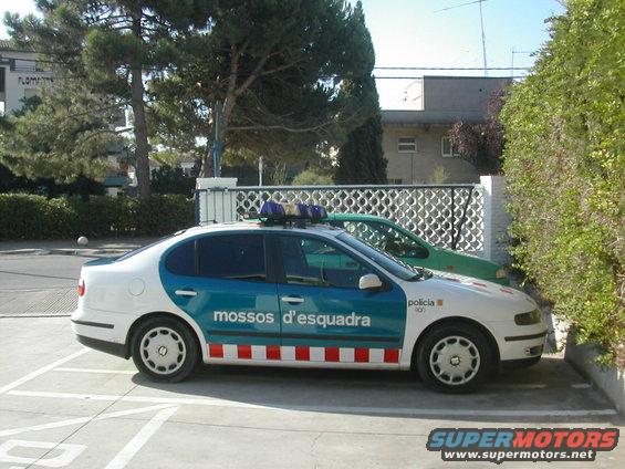 Side view of a white and blue police car with 'mossos d'esquadra' text, parked next to a green car