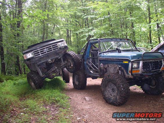 Modified blue 1979 Ford Bronco and black Ford truck off-roading in woods