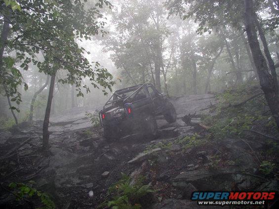 Off-road vehicle with roll cage climbing a rocky, wooded trail in fog