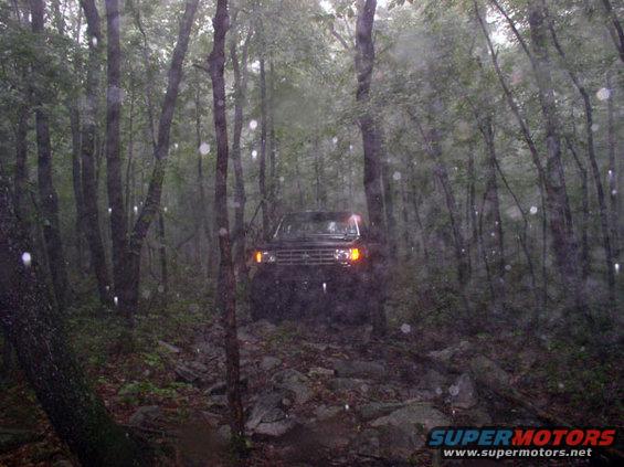 Black SUV with orange turn signals on a rocky trail, trees on either side, rain falling
