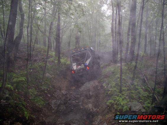 Off-road vehicle navigating a steep, muddy, wooded trail. It's tilted and kicking up dirt