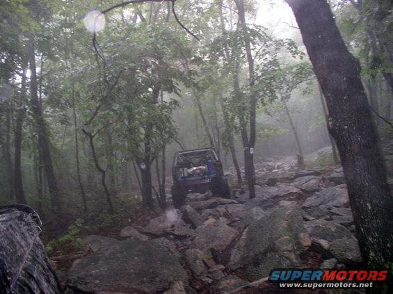 Blue off-road vehicle with roll cage navigating a rocky, wooded trail