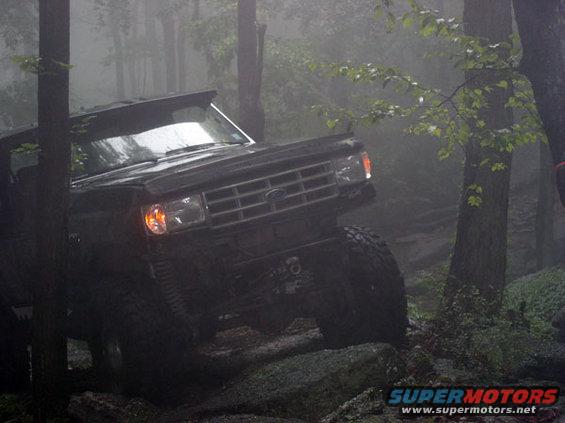 Black Ford Bronco navigating rocky terrain in foggy woods. Grille indicates 1987 model year