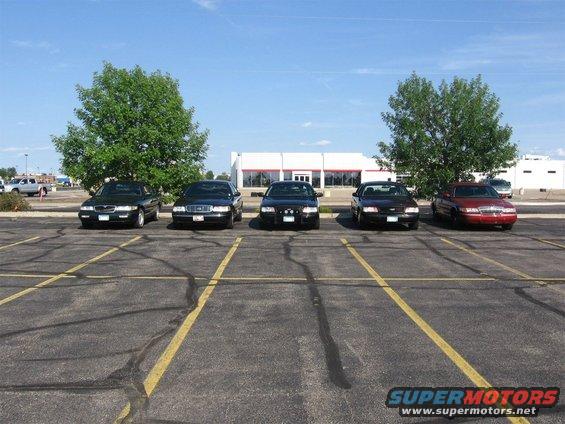 Five sedans parked in a lot: two dark sedans, two black sedans, and one red sedan. Building and trees in background