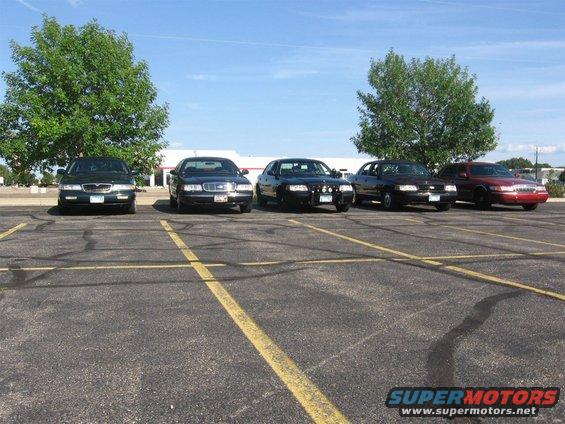 Five sedans parked in a lot, two dark green, three black, and one red. Trees and a building in the background