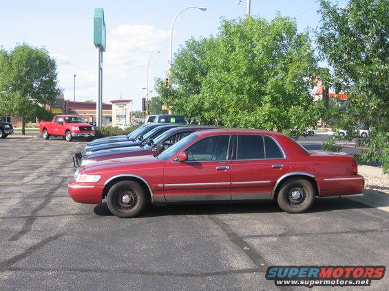 Red 1999 Ford Crown Victoria sedan, side view. Other cars and a red pickup truck are parked nearby