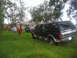 1995 Ford C-350 (Centurion Conversions) - A dark green and silver SUV is parked in a grassy yard near a damaged house, with people in red shirts working in the background.
