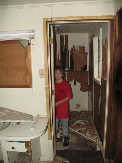 Boy in red shirt stands in doorway of room with damaged walls and debris on floor