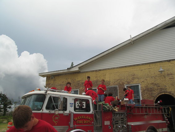 Red and white fire truck with "Waveland Fire Dept." text, children in red shirts on top