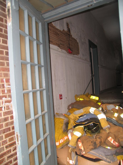 Pile of yellow and tan firefighter turnout gear with helmets and boots, inside a damaged building hallway
