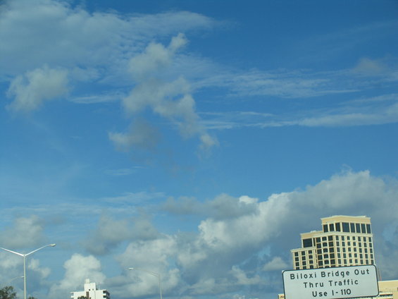 Blue sky with clouds over buildings and a sign that reads 'Biloxi Bridge Out Thru Traffic Use I-110'