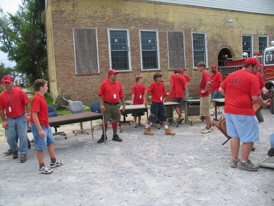 Group of people in red shirts standing around tables outside a brick building. A fire truck is visible on the right