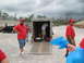 1995 Ford C-350 (Centurion Conversions) - Volunteers are seen unloading supplies from a trailer, with a black SUV parked nearby under a cloudy sky.