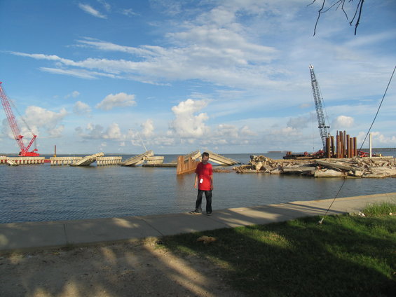 Person standing near a partially destroyed bridge with cranes and construction materials in the water