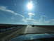 1995 Ford C-350 (Centurion Conversions) - A dark SUV travels down a highway that crosses over a body of water, with a bright sun and scattered clouds in the sky.