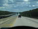 1995 Ford C-350 (Centurion Conversions) - A dark SUV travels down a highway bridge that spans over a body of water, flanked by dense trees under a cloudy sky.