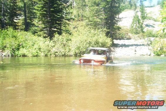 Jeep driving through a river with trees and a rocky bank in the background