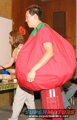 Person in a red tomato costume with green collar, standing indoors with others
