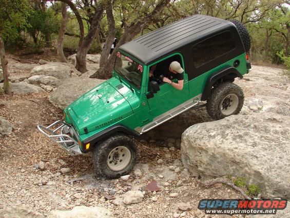 Green Jeep Wrangler Rubicon off-roading over rocks, with a black hardtop and spare tire on the back