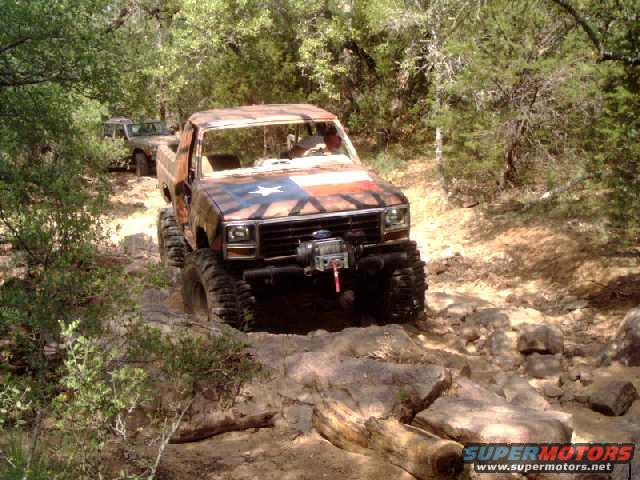 Modified pickup truck with Texas flag paint on hood, driving on rocky trail. Another SUV follows behind