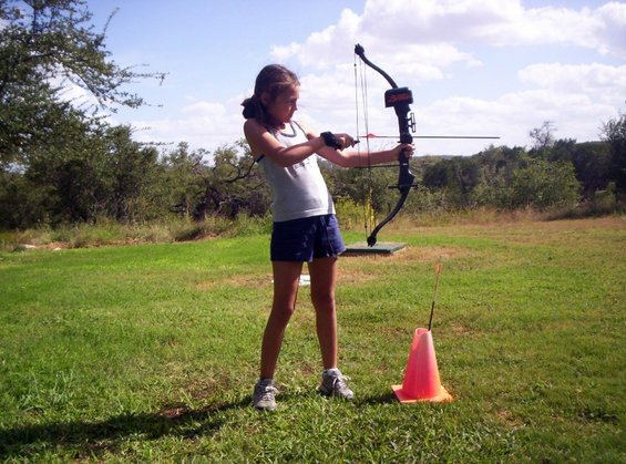 Girl with bow and arrow drawn, aiming at a target in a grassy field